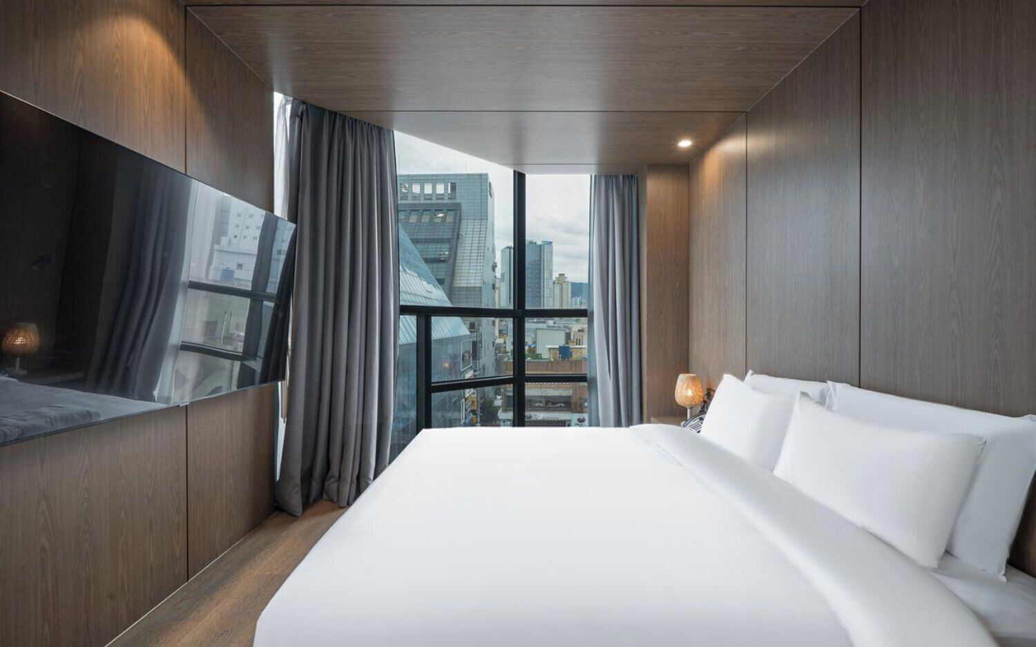 A hotel room with a large bed, a flat-screen TV, and a city view through the window.