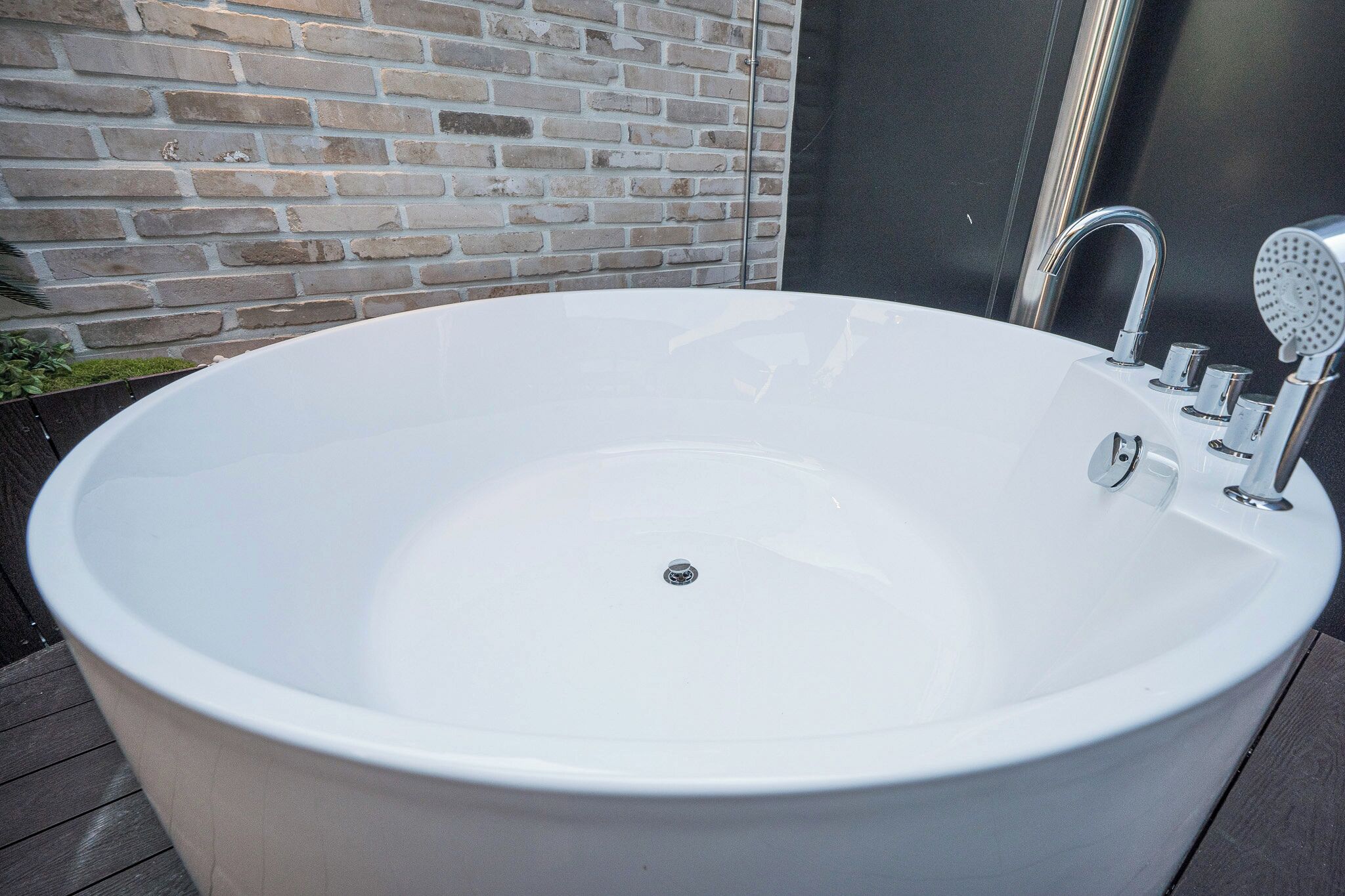Une grande baignoire blanche, autoportante, dotée d’un robinet et d’une pomme de douche au design moderne, placée devant un mur de briques.