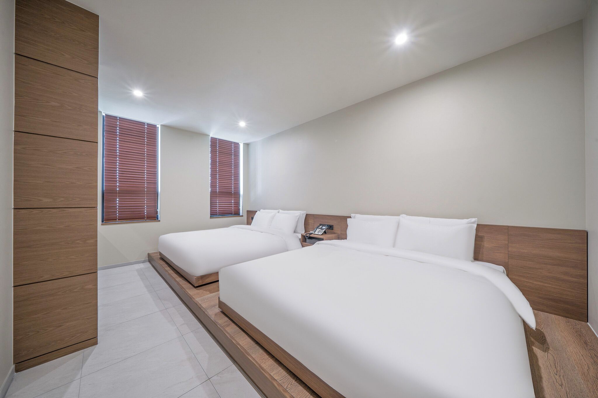 A modern hotel room with two beds, wooden headboards, and a large window with blinds.