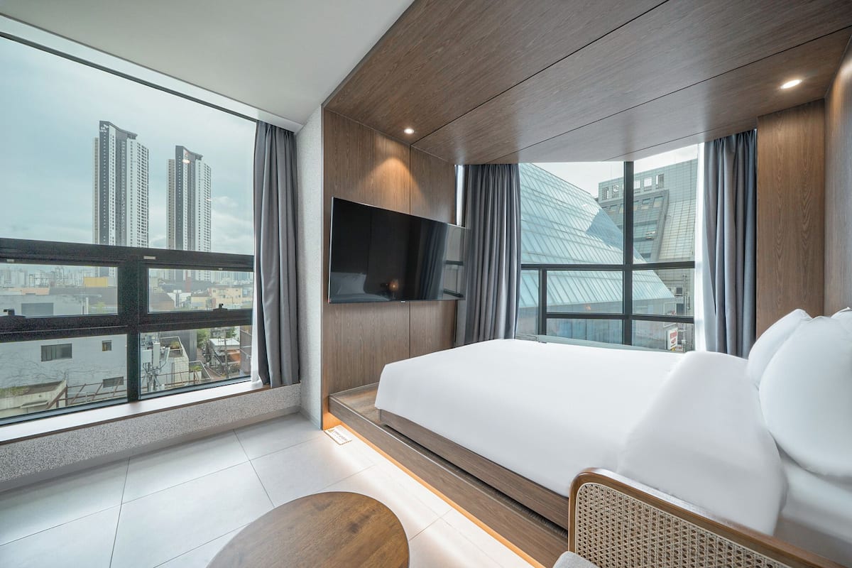 A modern hotel room with a large bed, a flat-screen TV mounted on the wall, and a view of city buildings through large windows.