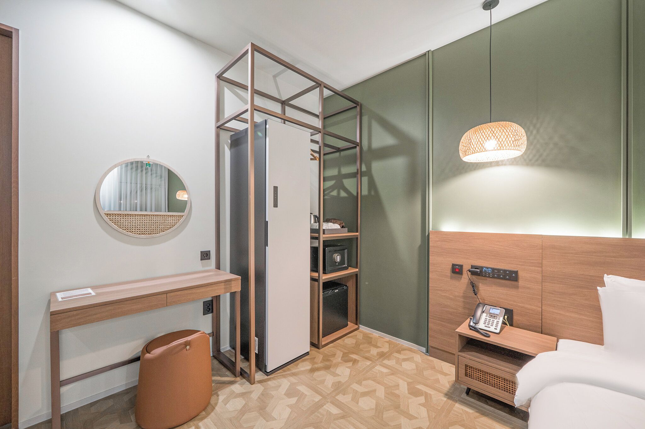 A compact hotel room with a bed, a desk, a wardrobe, a mirror, and a small trash bin.
