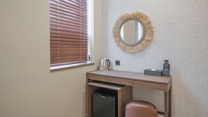 A compact room with a wooden desk, a small refrigerator, a mirror, and a window with blinds. - Giant Hotel (Daegu)