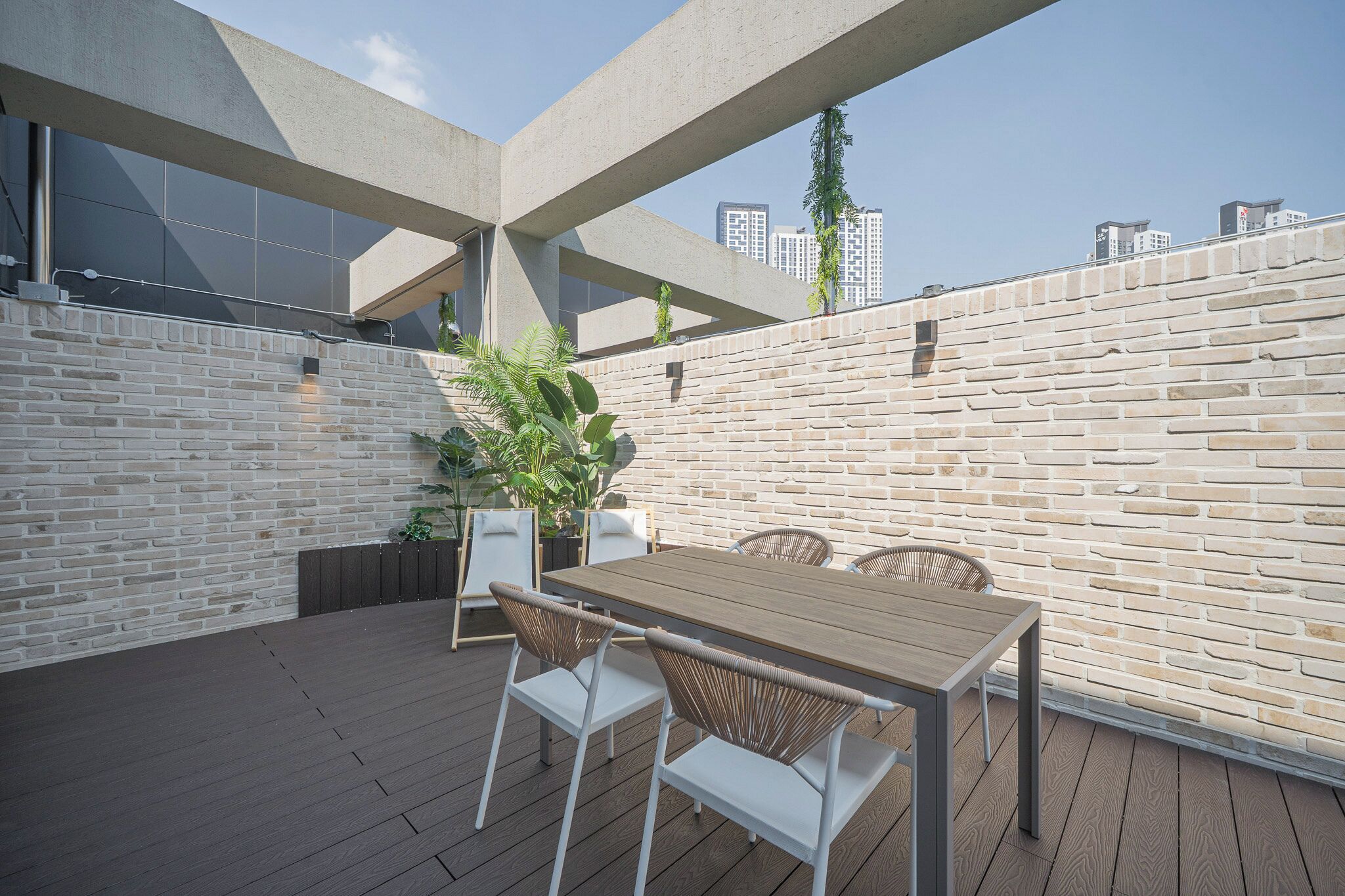 Een terras buiten met een houten tafel en witte stoelen, omgeven door een bakstenen muur en een glazen balustrade.