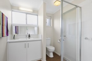 Un baño moderno con lavamanos, espejo y retrete blancos. Cuenta con una zona de ducha con puerta de cristal.