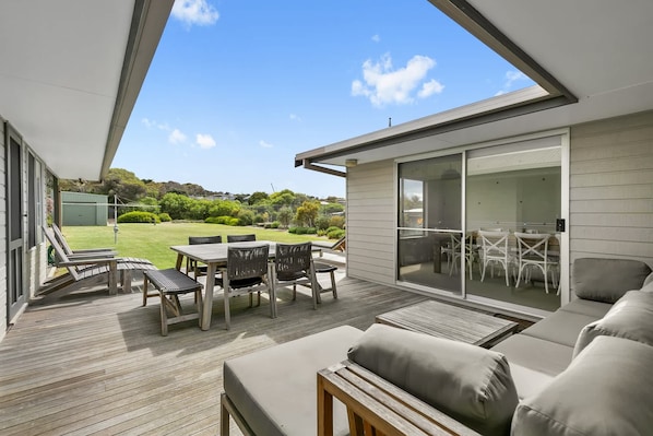 Un patio moderno con cubierta de madera, mobiliario exterior y vistas a un jardín exuberante y colinas al fondo.