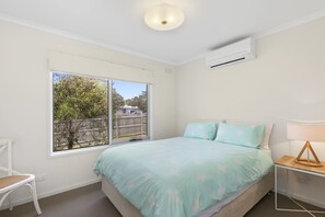 Un dormitorio con cama, una silla, mesita de noche, lámpara y ventana con vista a una cerca y árboles.