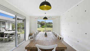 A dining area with a wooden table and white chairs, a black and yellow pendant light, and a view of a garden through large glass doors.