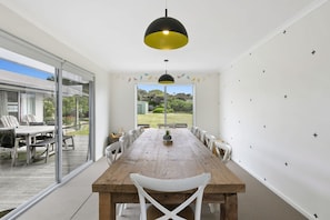 Un comedor con mesa de madera y sillas blancas, una lámpara colgante negra y amarilla, y vistas a un jardín a través de amplios ventanales.