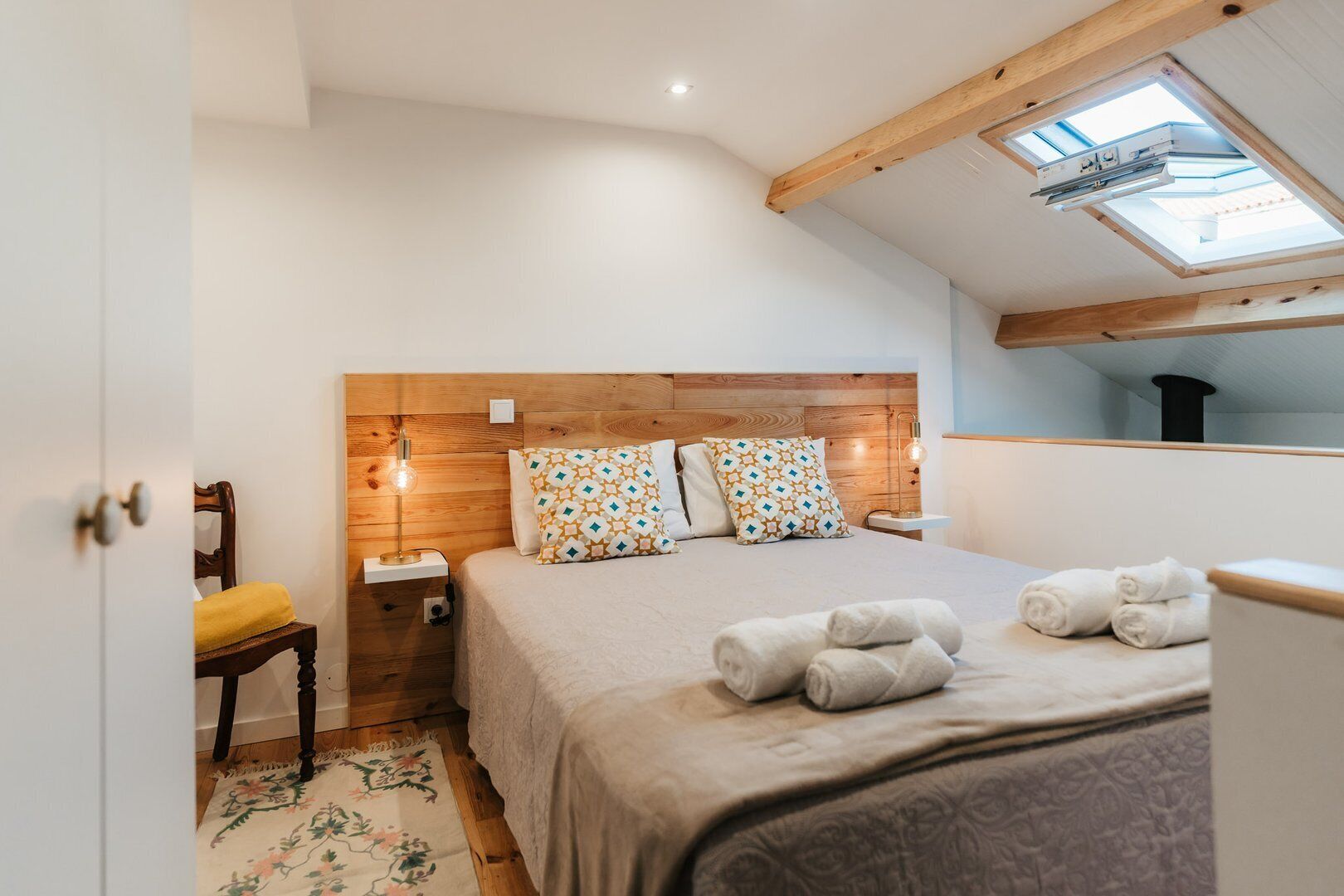 Un dormitorio con cabecera de madera, almohadas estampadas, una mesita de noche con lámpara y un tragaluz sobre la cama.