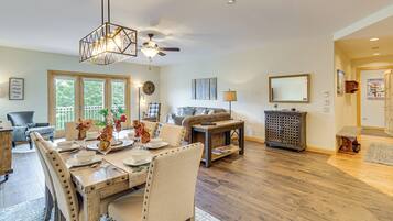 A spacious living room with a wooden dining table, comfortable seating, a ceiling fan, and a large window offering a view of the outdoors.