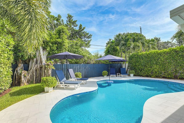 A swimming pool with lounge chairs and umbrellas, surrounded by greenery and a wooden fence. - Private Pool + Backyard: Villa in North Palm Beach (North Palm Beach)