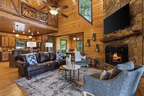 A cozy cabin interior with a stone fireplace, wooden walls, a leather sofa, and a television mounted on the wall. - EZ Come EZ Geaux - Stunning Mtn Views - Hot Tub (Blue Ridge)