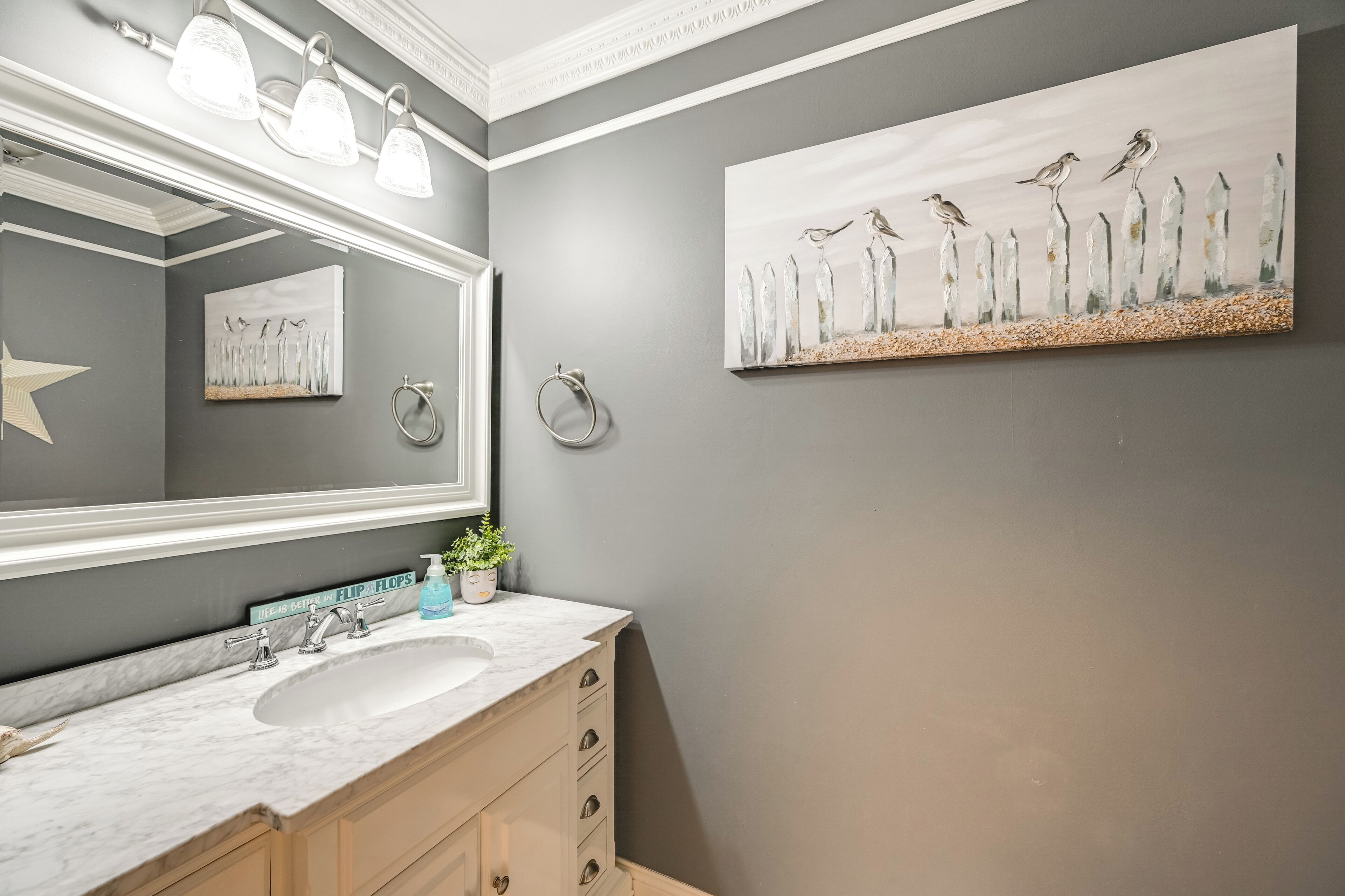 Un bagno con uno specchio grande, un lavandino e un portasciugamani. Al muro c'è un quadro e sul ripiano un piccolo vaso di fiori.