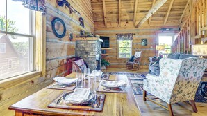 A wooden cabin interior with a dining table set for four, a floral armchair, and a stone fireplace. - 2 Mi to Mabry Mill: Mtn-View Cabin w/ Porch! (Meadows of Dan)