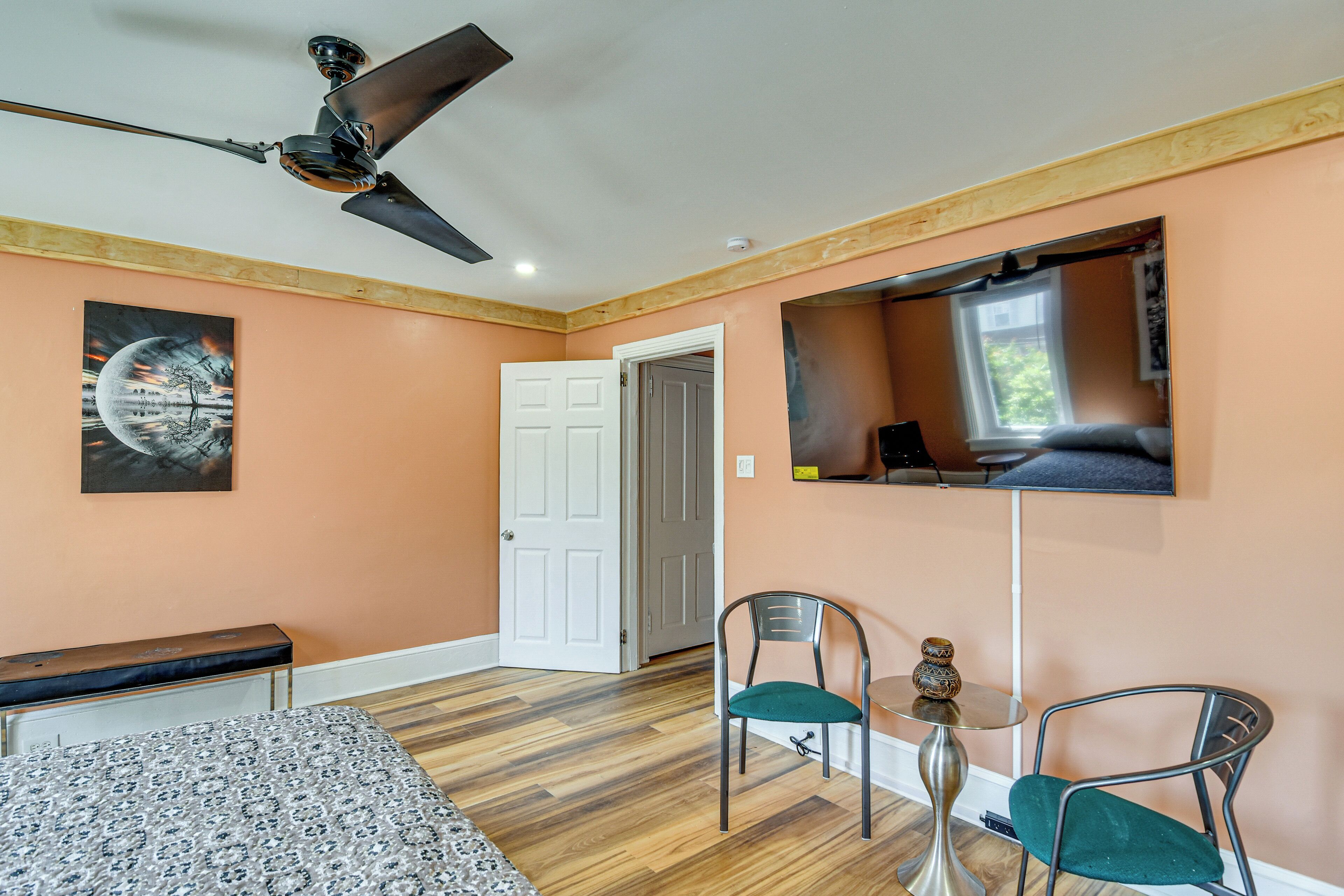 A bedroom with a ceiling fan, a bed, a bench, two chairs, a small table, and a TV mounted on the wall.