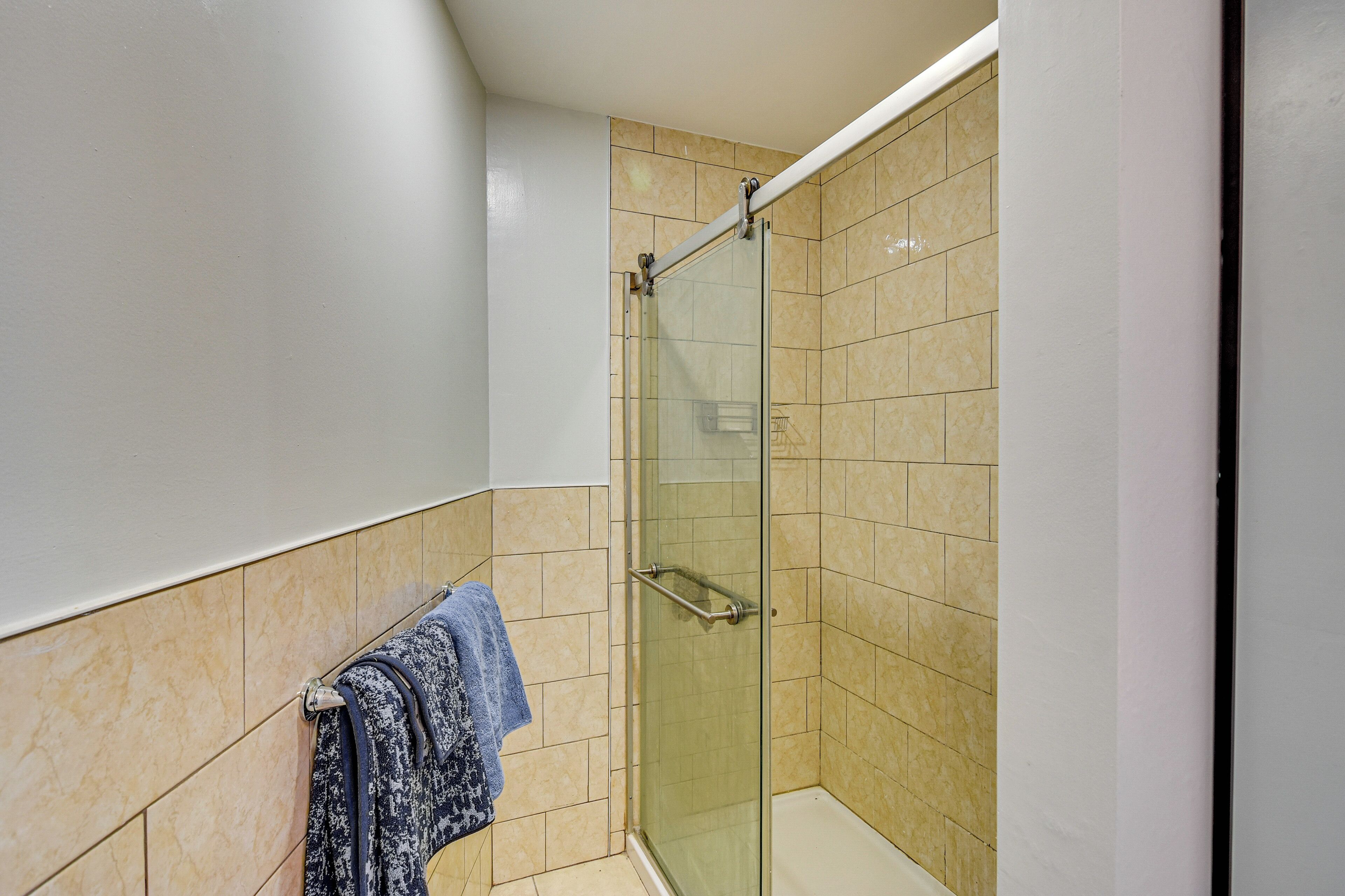 A corner bathroom with a glass shower enclosure, beige tiles, and a towel rack with a blue towel.