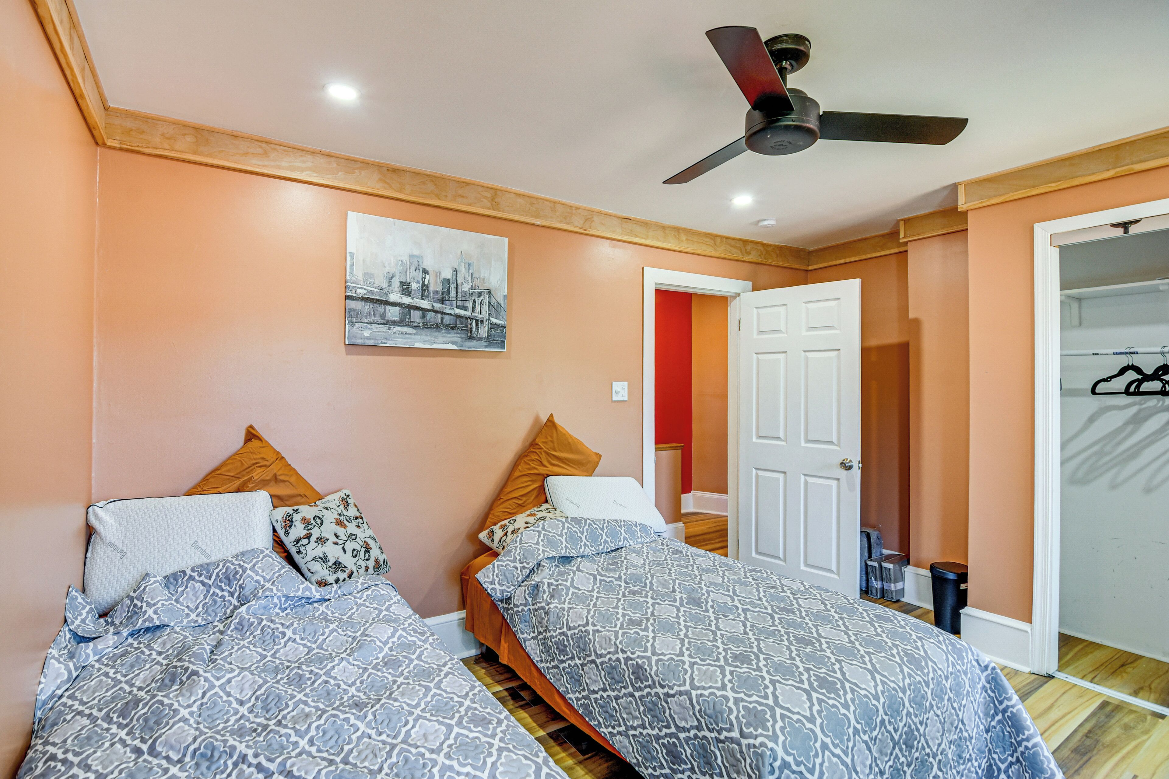 A bedroom with two beds, a ceiling fan, a painting on the wall, and a door leading to another room.