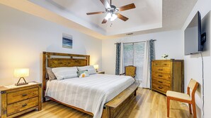 Une chambre à coucher avec un lit en bois, une table de chevet, un banc, une chaise, un ventilateur de plafond et un téléviseur fixé au mur.