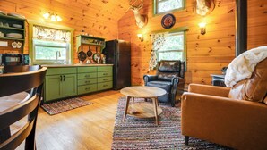A cozy cabin interior with wooden walls, a dining area with a table and chairs, a kitchen with green cabinets, and a fireplace with a deer head mounted above it. - On 30 Acres w/ Trails & Garden: Stockholm Cabin! (Stockholm)