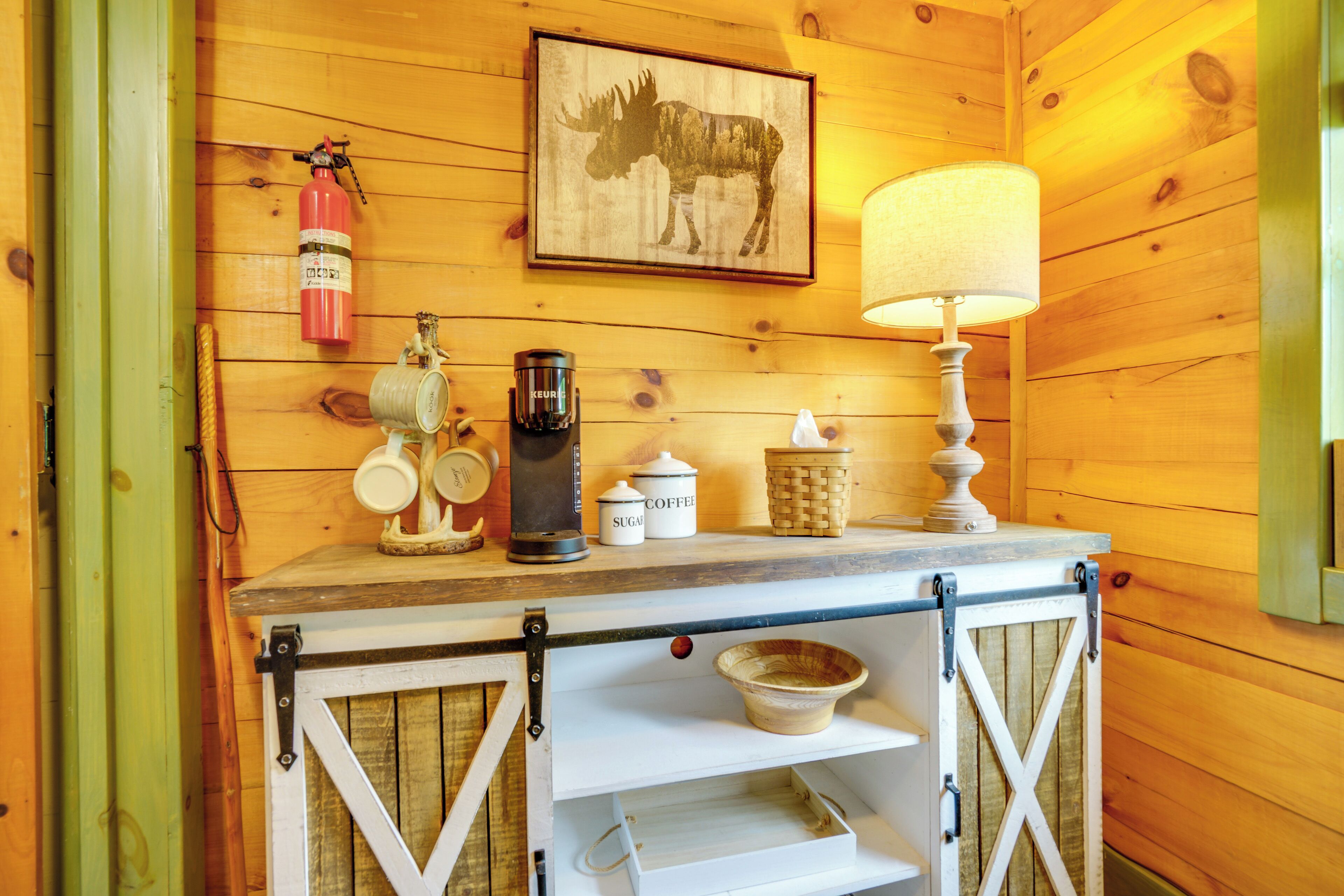A wooden interior with a coffee station, a framed moose picture, a fire extinguisher, and a lamp.