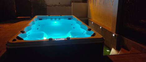 Un bain à remous avec un éclairage bleu, entouré d’un mur de pierres et d’une terrasse en bois.
