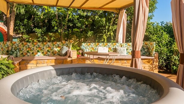 A hot tub with a stone surround, a covered seating area with a floral pattern, and a stone wall with greenery in the background. - 15 minutes by car / Sitges – Villa Ilus. XXL Pool, A/C by Weekly Villas 12 P (Sitges)