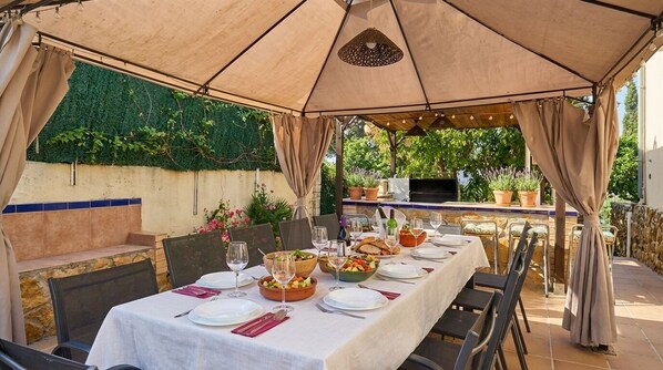 Un área de comedor al aire libre con una mesa lista para comer, rodeada de sillas y un toldo arriba.