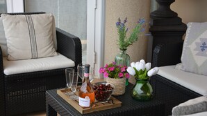 Un sofá de mimbre con un cojín, una mesita con una botella de vino, copas, un bol de cerezas y un jarrón con flores.