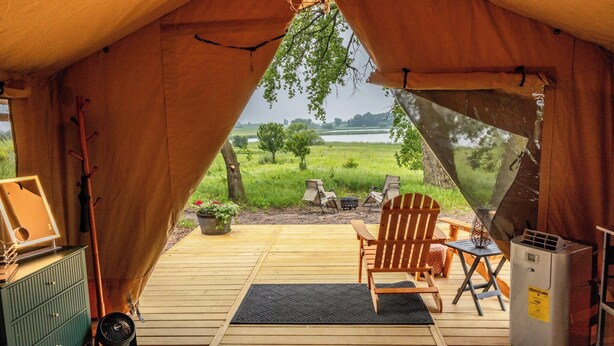 Een tent met uitzicht op een grasveld en een meer. Binnenin staat een houten stoel, een klein tafeltje en een heater.