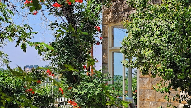 A stone building with a glass door, surrounded by lush greenery and red flowers. - 4-bedroom Villa with Heating, Pool, Playgrounds, WiFi, Gardens: Douq, Batroun (محافظة الشمال)