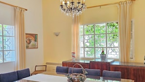 A dining area with a glass table, dark chairs, a chandelier, and a view of greenery through large windows. - 4-bedroom Villa with Heating, Pool, Playgrounds, WiFi, Gardens: Douq, Batroun (محافظة الشمال)