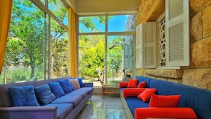 A spacious living area with large windows, a stone wall, and a mix of blue and red sofas. - 4-bedroom Villa with Heating, Pool, Playgrounds, WiFi, Gardens: Douq, Batroun (محافظة الشمال)