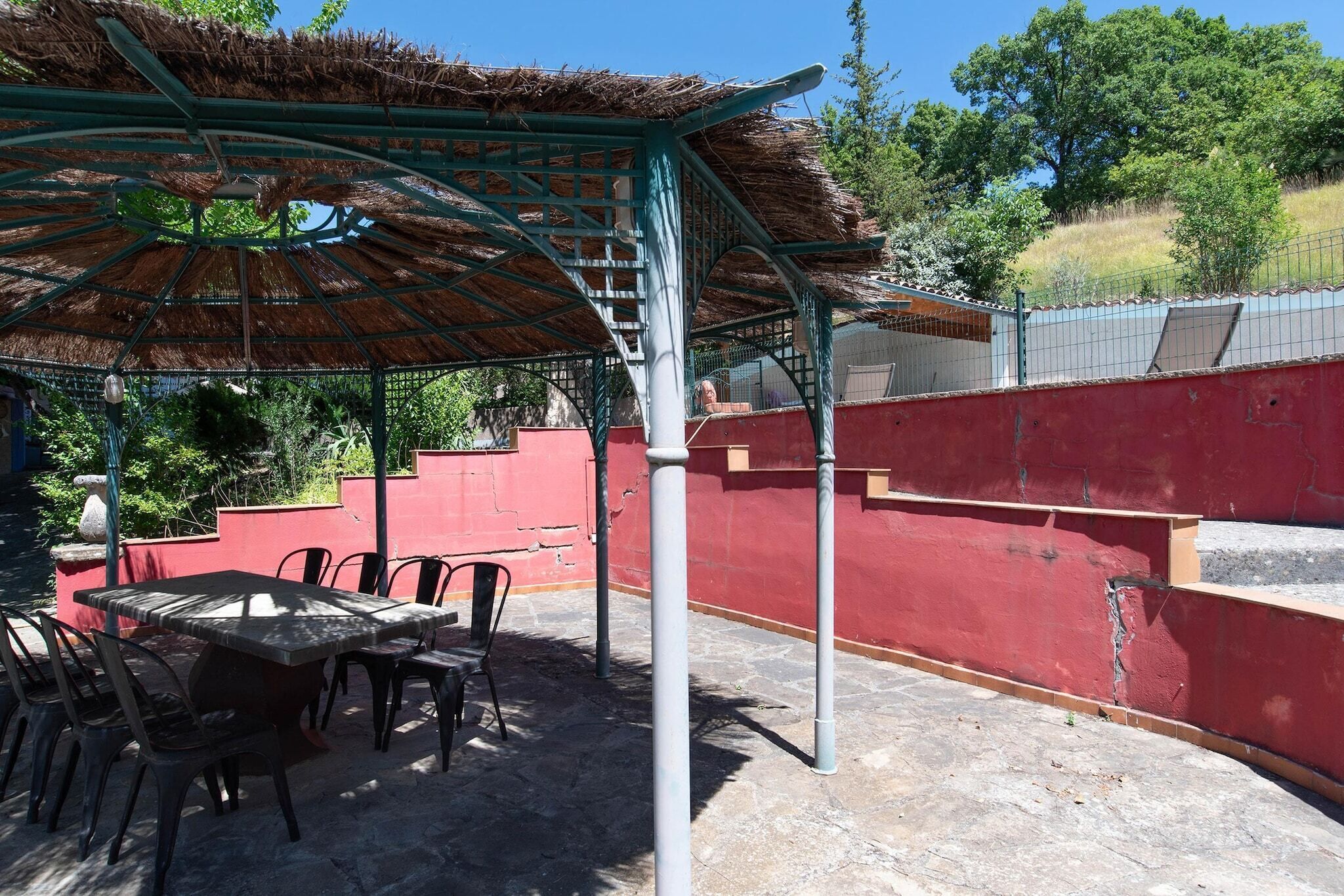 A covered outdoor seating area with a table and chairs, a thatched roof, and a red wall.