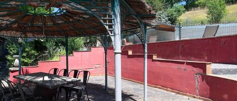 A covered outdoor seating area with a table and chairs, a thatched roof, and a red wall.