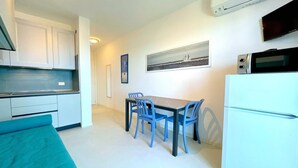 A compact kitchen with white cabinets, a dining table with blue chairs, a white refrigerator, and a flat-screen TV mounted on the wall.