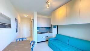 A compact living space with a dining table, blue chairs, a turquoise sofa, a kitchenette, and a framed picture on the wall.