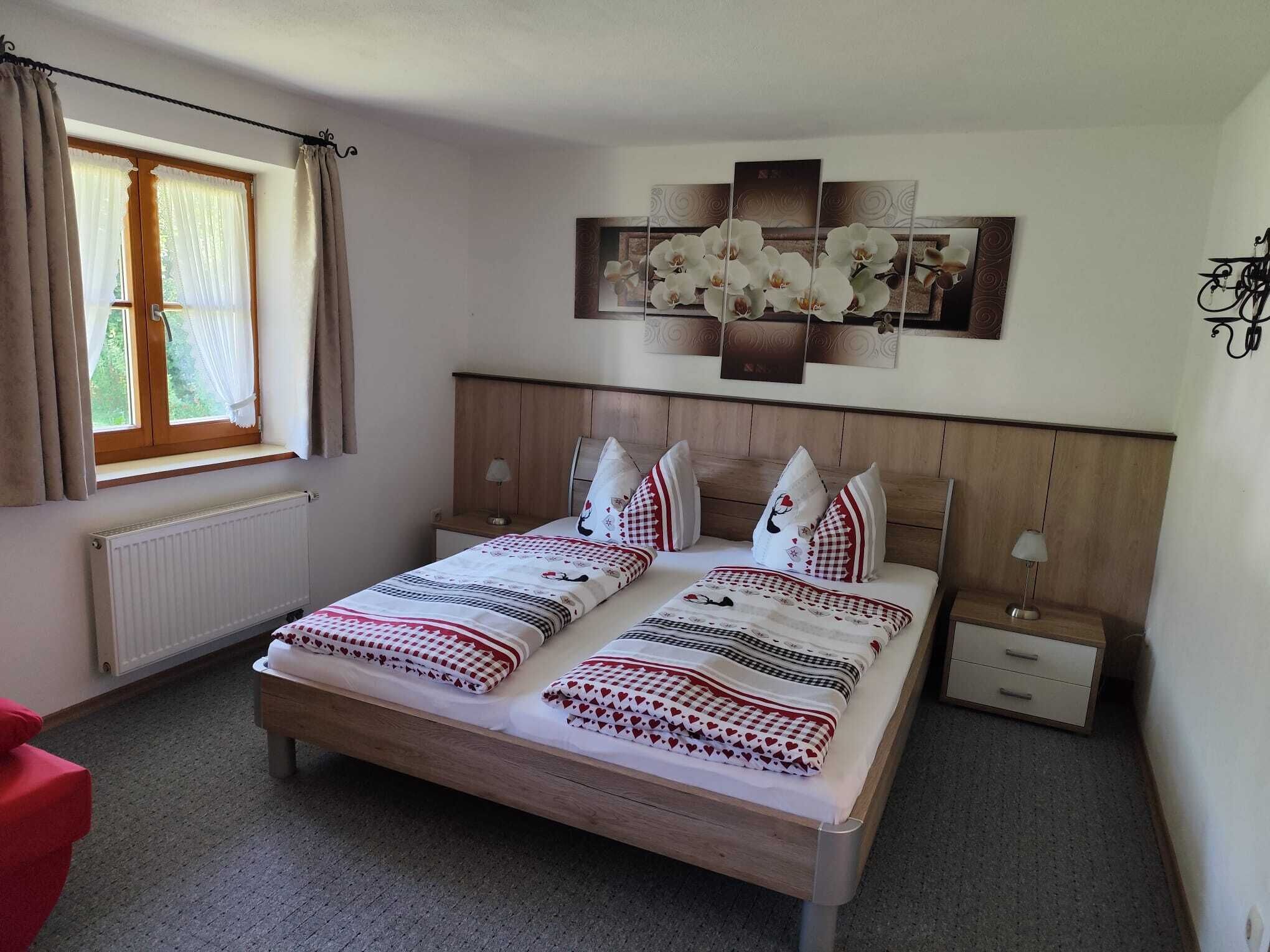 Ein Schlafzimmer mit einem hölzernen Bett, zwei Nachttischen, einer Heizung, einem Fenster mit Vorhängen und einem an der Wand hängenden Kunstwerk.