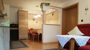 Un salon chaleureux avec un canapé rouge, une petite table recouverte d’une nappe blanche, une table à manger en bois avec des chaises, et un coin cuisine équipé d’un four à micro-ondes.