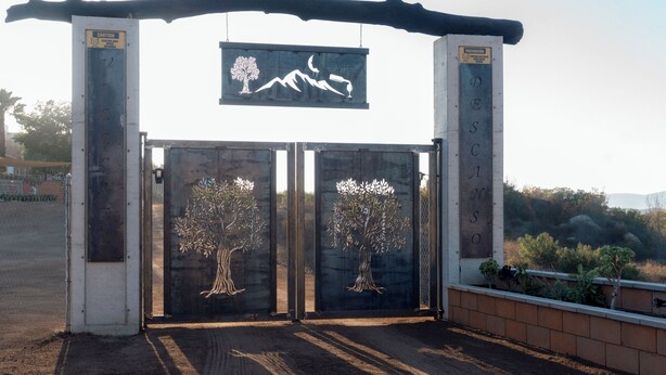 Una entrada con portón y un letrero que muestra un árbol y montañas, además de una placa con la palabra 'DESCANSO'.