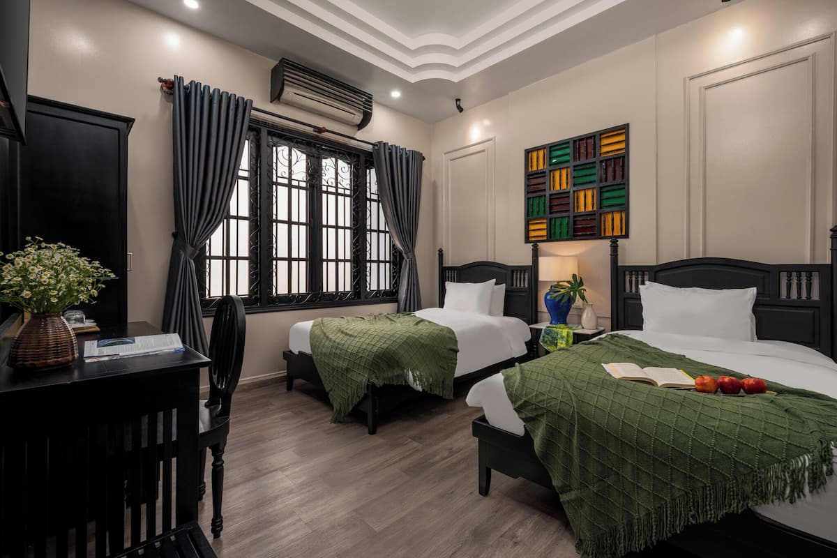 Ein Hotelzimmer mit zwei Betten, einem Schreibtisch mit Stuhl, einem Fenster mit Vorhängen, einer Klimaanlage und einem farbenfrohen Kunstwerk an der Wand.