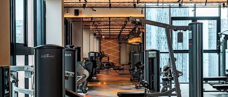 A modern gym with various exercise machines, large windows, and a geometric light fixture.