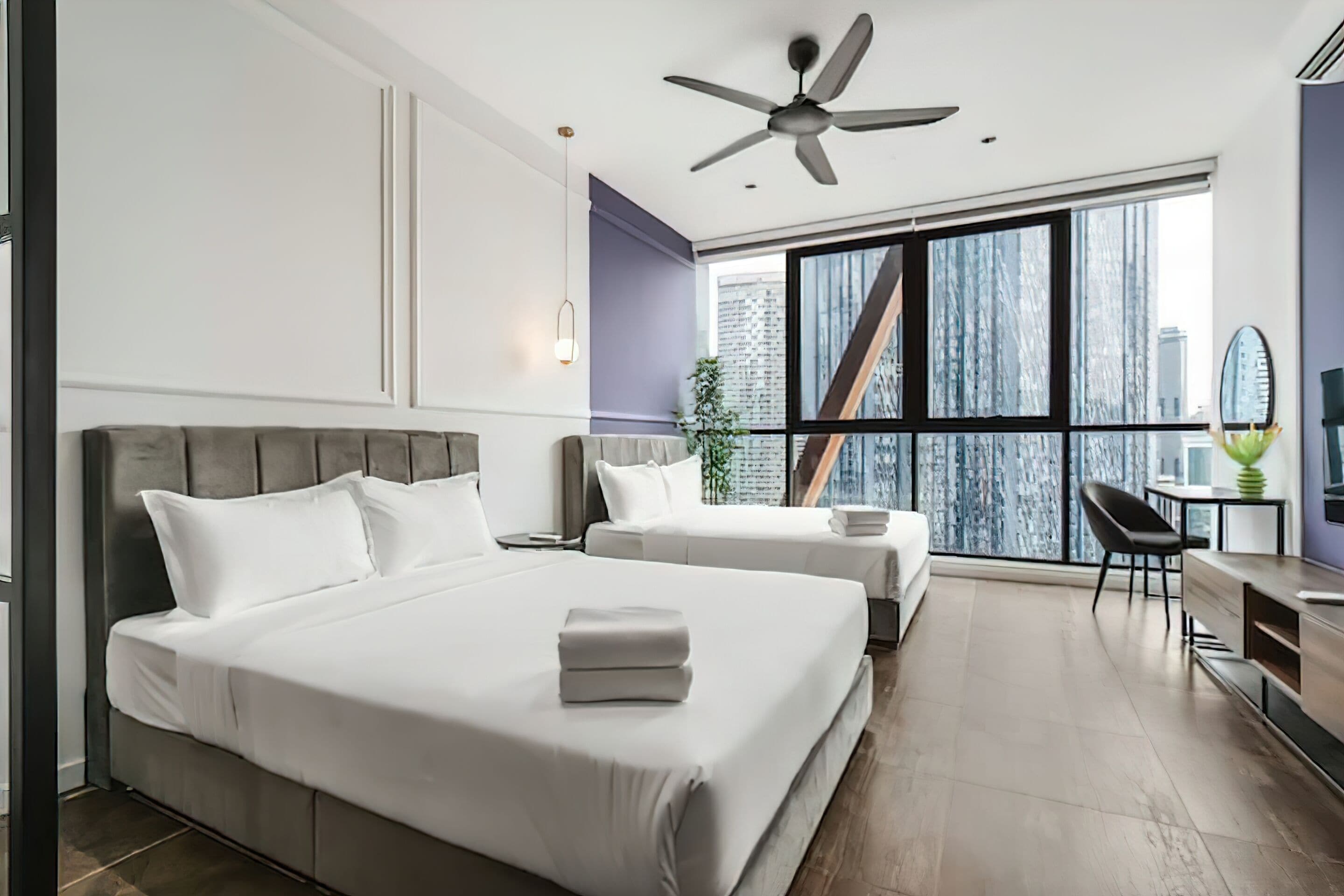 Ein modernes Hotelzimmer mit zwei Betten, einem Deckenventilator, einem großen Fenster mit Blick auf die Stadt, einem Schreibtisch mit Stuhl und einer kleinen Pflanze.