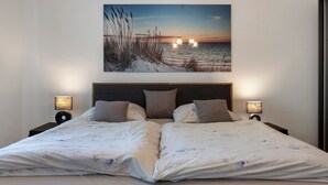 A double bed with floral bedding, two bedside lamps, and a sunset beach painting above. - Apartment 'Am Herforder Tor Für' with Private Garden, Balcony and Wi-Fi (Bad Salzuflen)