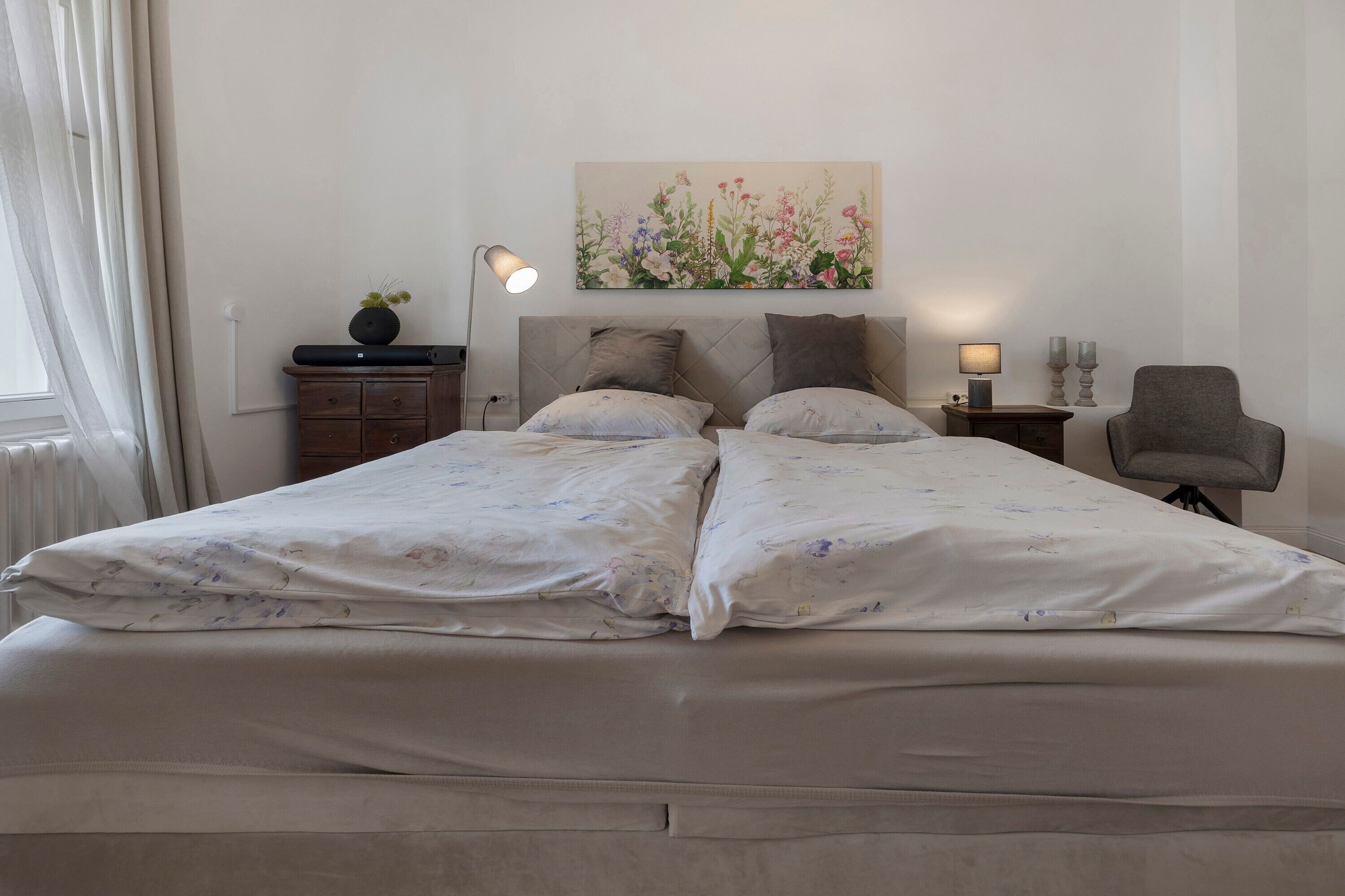 A double bed with floral bedding, a bedside table with a lamp, a small cabinet, and a chair.