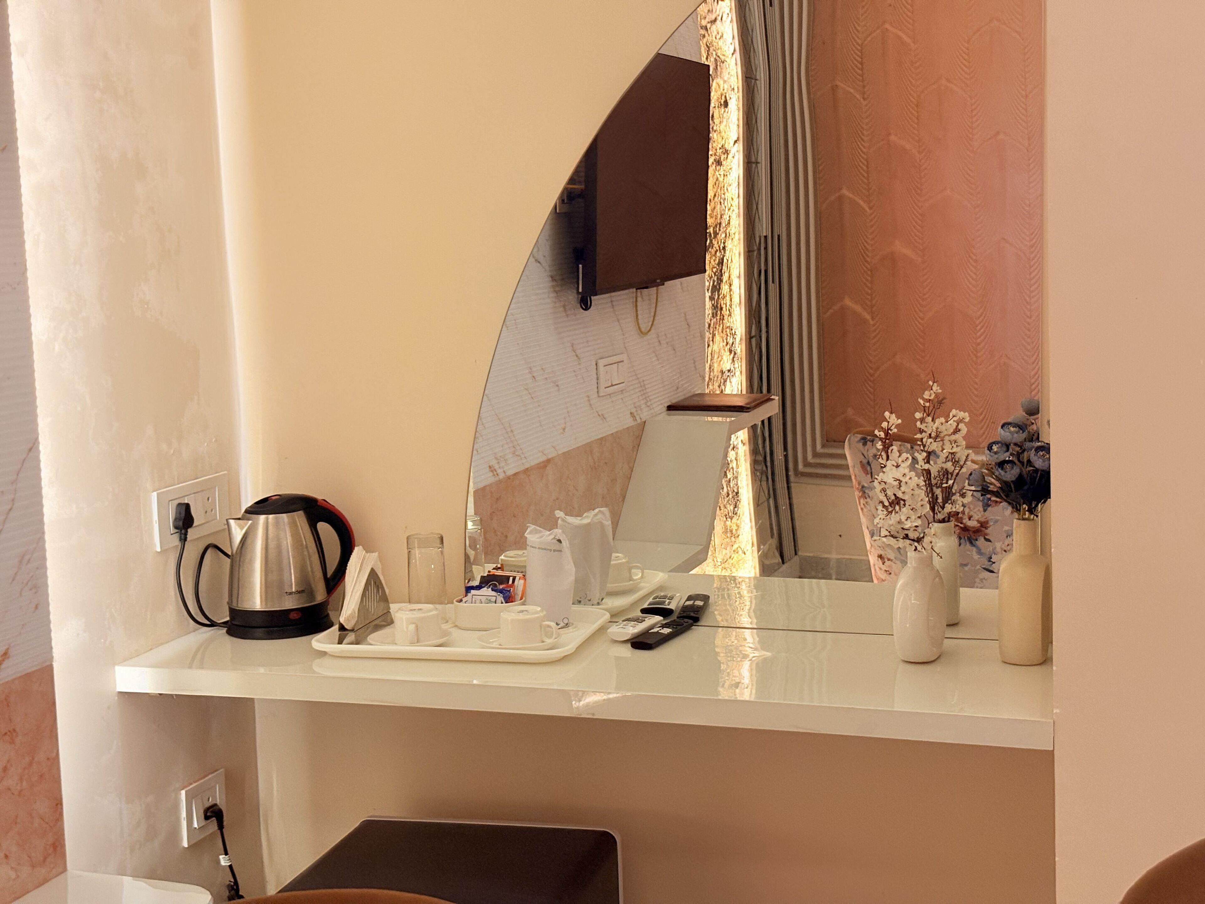 Camera d'albergo con un ripiano bianco, una macchina per il caffè, un vassoio con tazze e un telecomando, e un vaso con fiori.