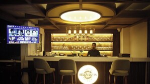 A bar area with a bartender, a TV screen displaying movie options, and a circular illuminated sign reading "BELLUJA". - Hotel Q Green (Nalbari)