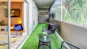 A narrow balcony with artificial grass, a glass door leading to a room with a wooden panel wall, and a large window with a tropical view. - Cordova Arms by Rocketstay (Fort Lauderdale)
