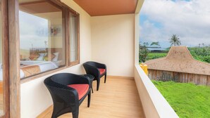 A balcony with wicker chairs, a bed with white bedding, and a view of a thatched-roof building and greenery. - Jnana Bali Villa (Sukawati)