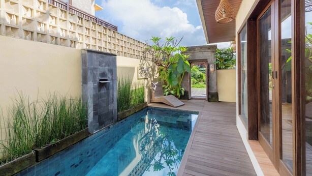 A swimming pool with a wooden deck, a stone wall, and a pergola with plants. - Jnana Bali Villa (Sukawati)