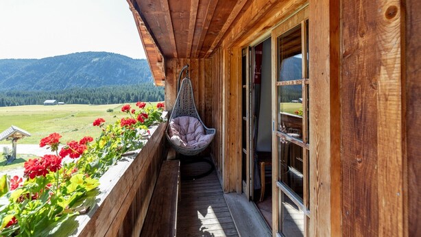 Un balcon en bois agrémenté d’un fauteuil suspendu, de balustrades en bois et offrant une vue sur un paysage verdoyant avec des montagnes.